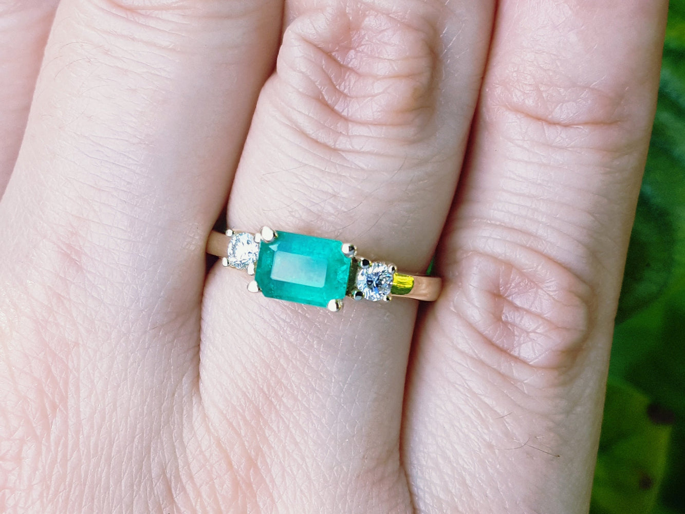Yellow gold emerald ring on a person's finger against a blurred natural background.