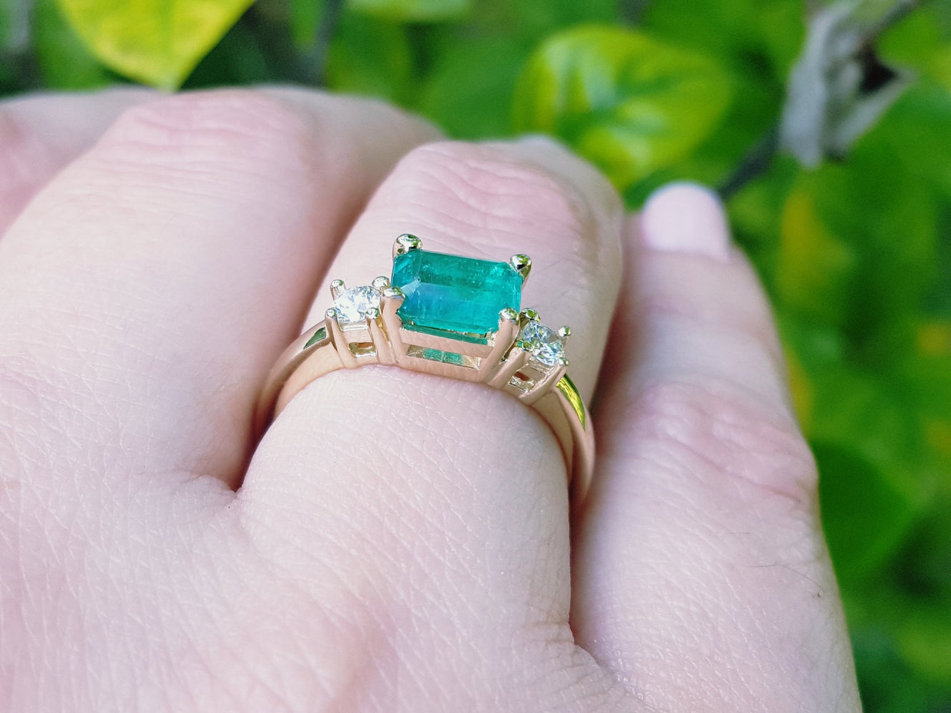 A yellow gold emerald ring three stone on a hand against a blurred green background