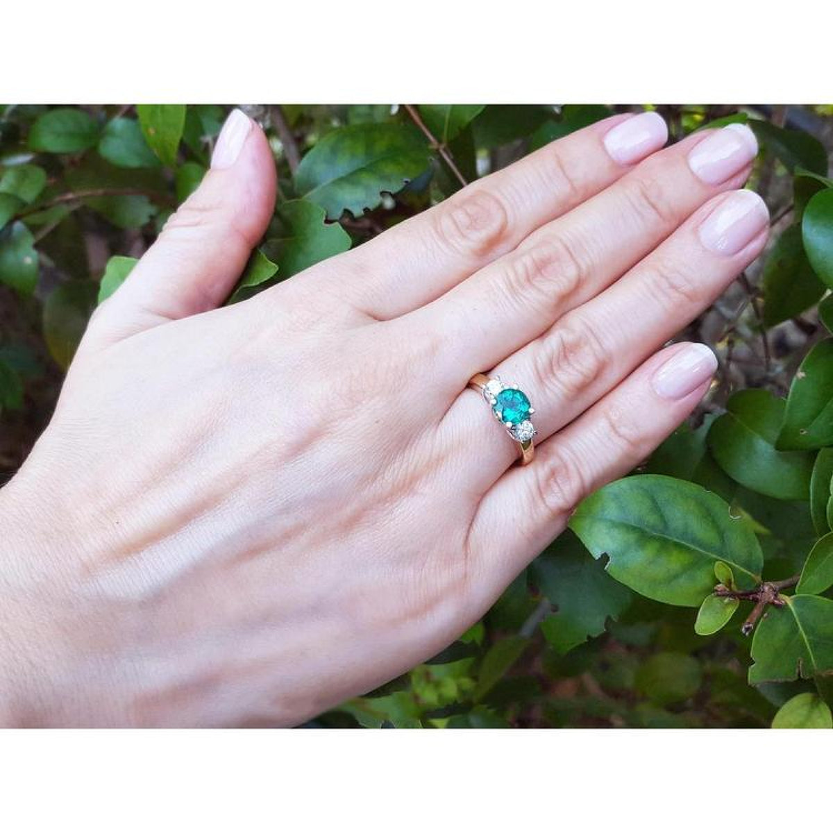 14k yellow gold three-stone ring with a round bluish-green emerald and two side diamonds on a hand.