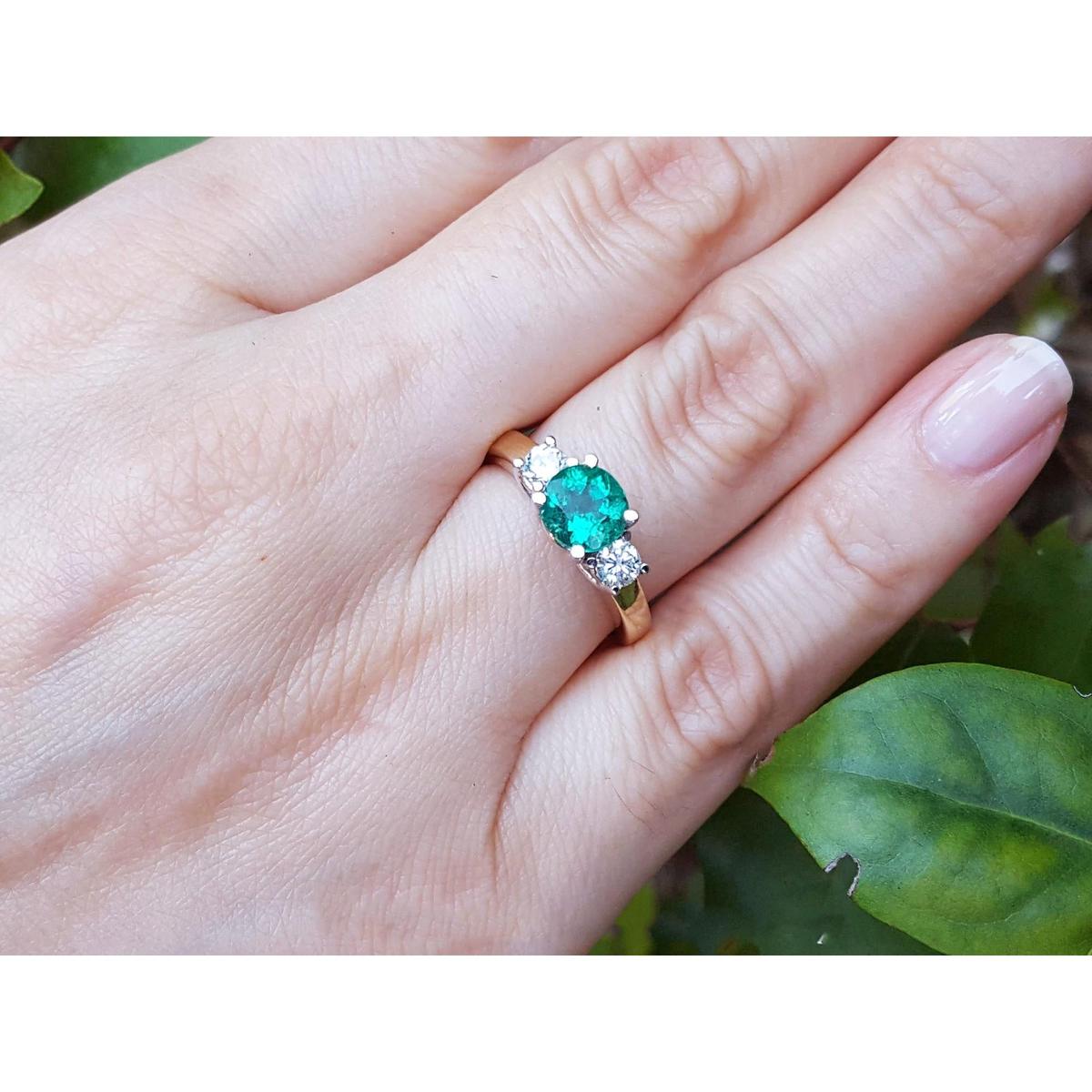 Ring with a round emerald and side diamonds on a hand against a leafy background.
