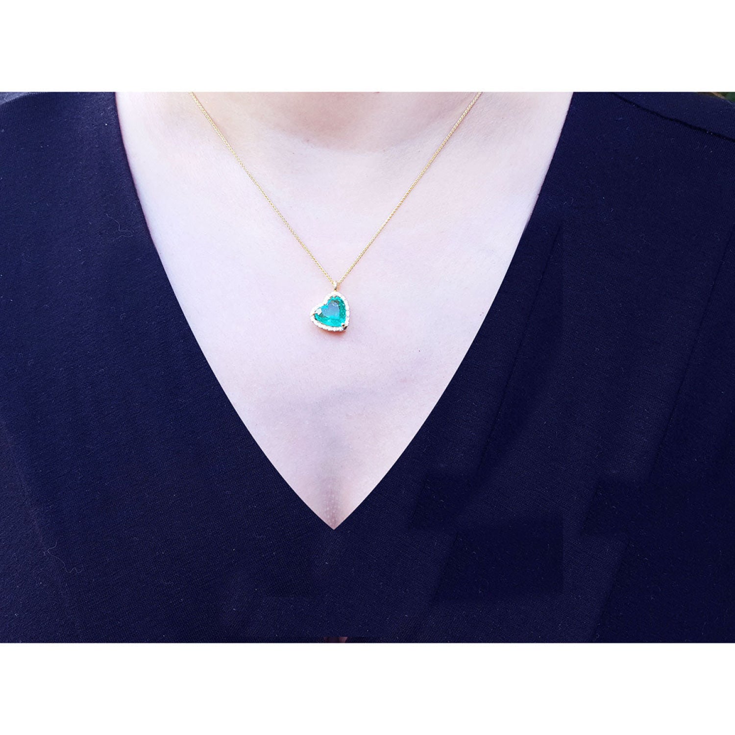Tilted heart emerald necklace with halo diamonds on an 18K gold chain, worn on a woman's neck.