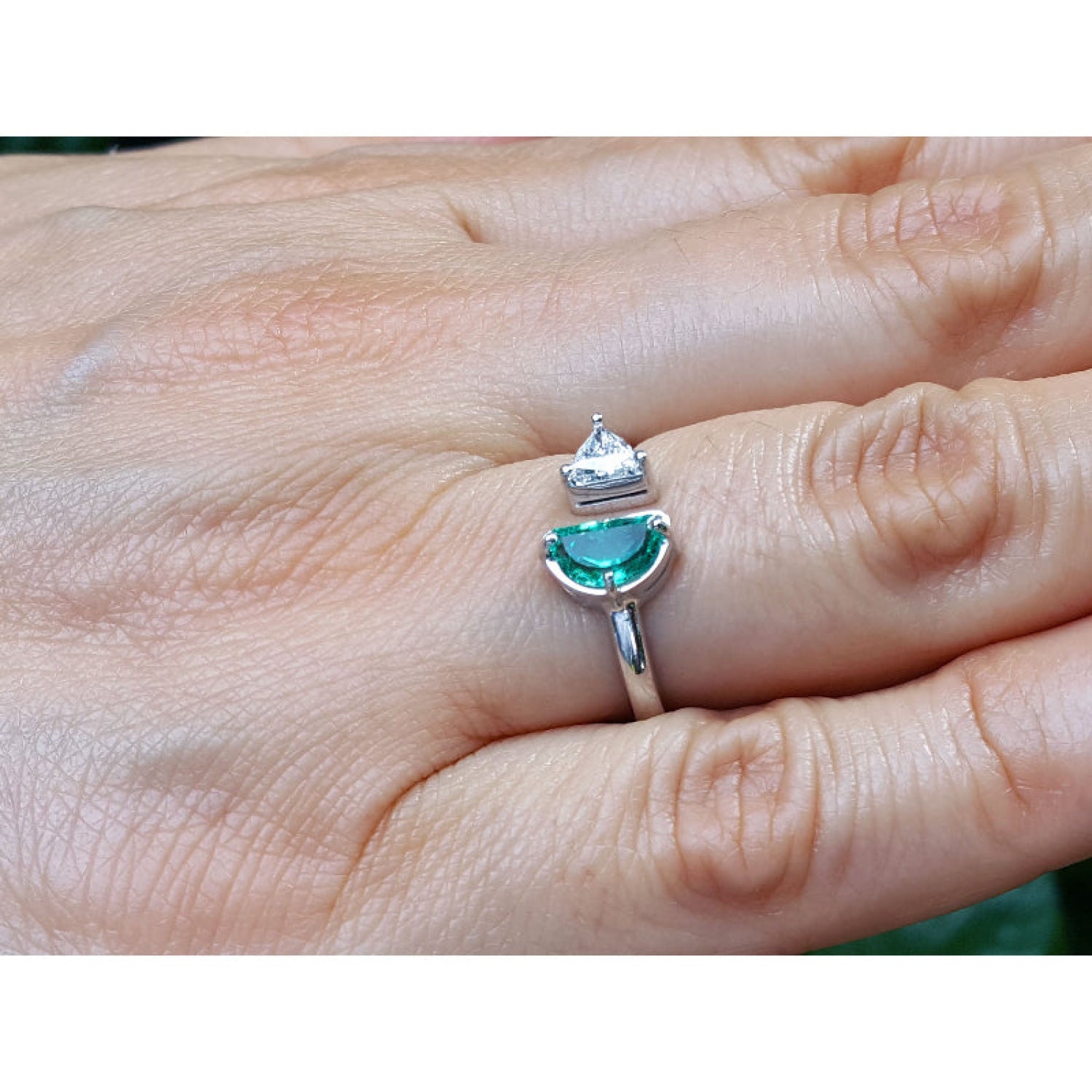 A negative space Toi et Moi ring features a half-moon-shaped natural Colombian emerald and a trillion-cut diamond, on a person's finger.