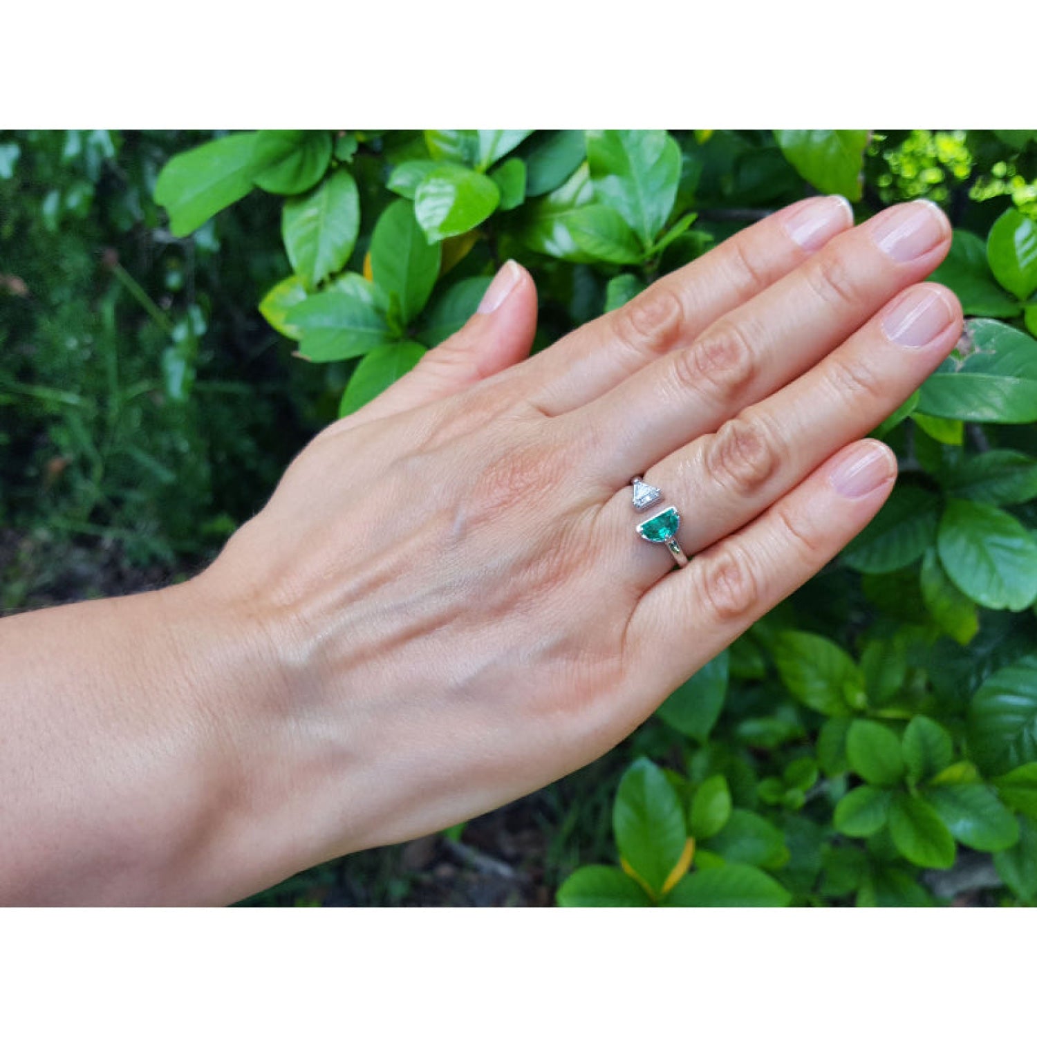 A 18k white gold toi et moi ring with a half-moon natural colombian emerald worn on a person's ring finger, shown against a background of green outdoor foliage.