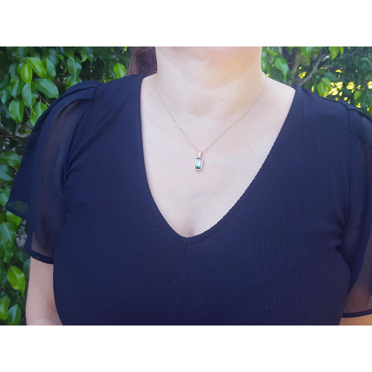 Person wearing a dark blue top with a tourmaline necklace against a green leafy background.