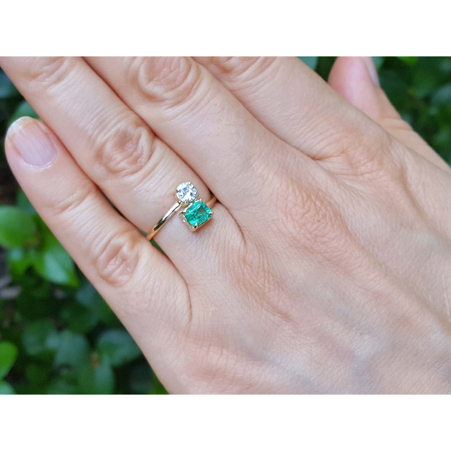 A close-up of a hand wearing a You and Me 18k gold ring with a natural emerald and diamond, set against a blurred green outdoor background.