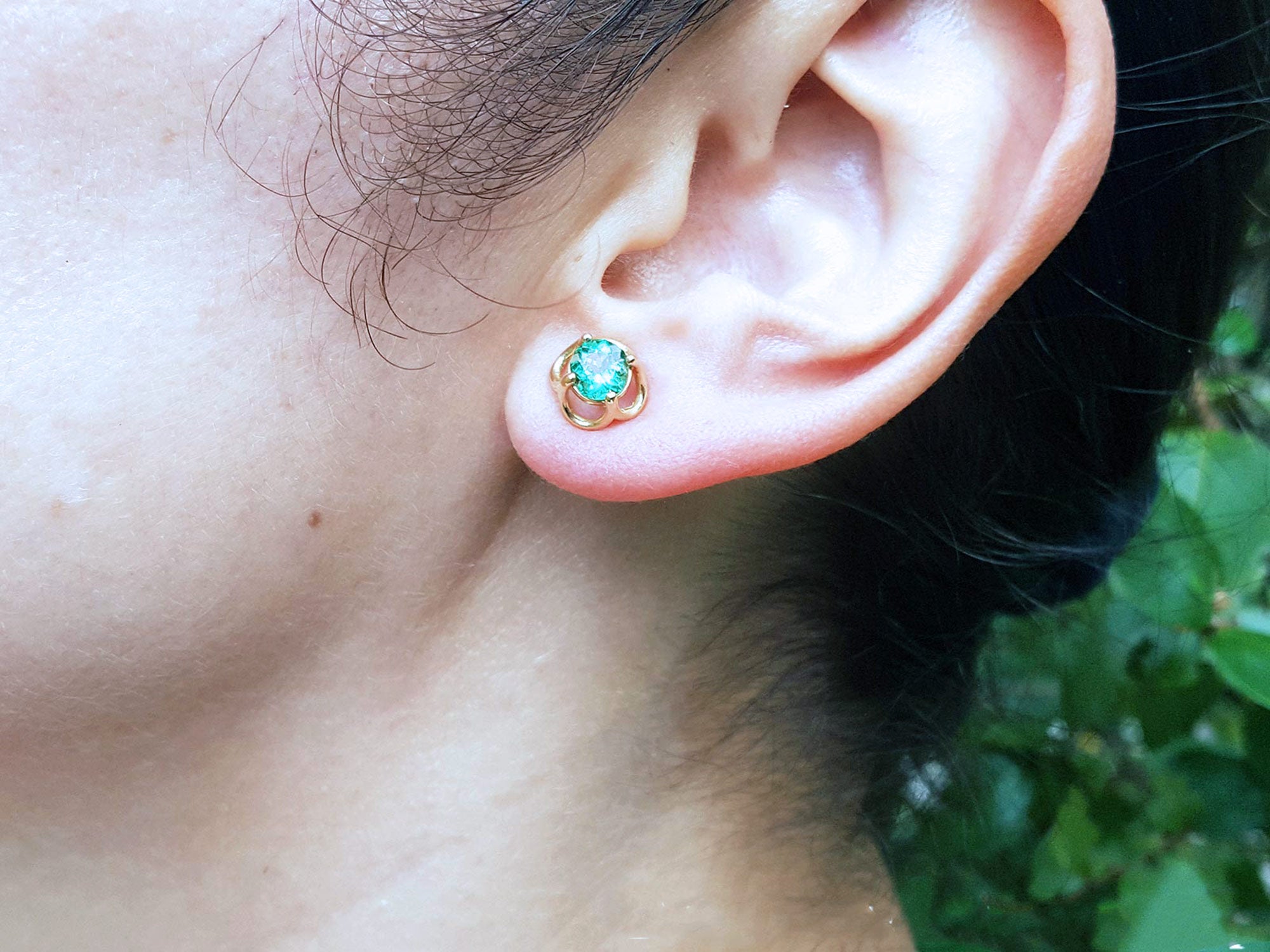 Close-up of a woman&#39;s ear wearing a natural tulip emerald stud earrings in a blurred natural background.