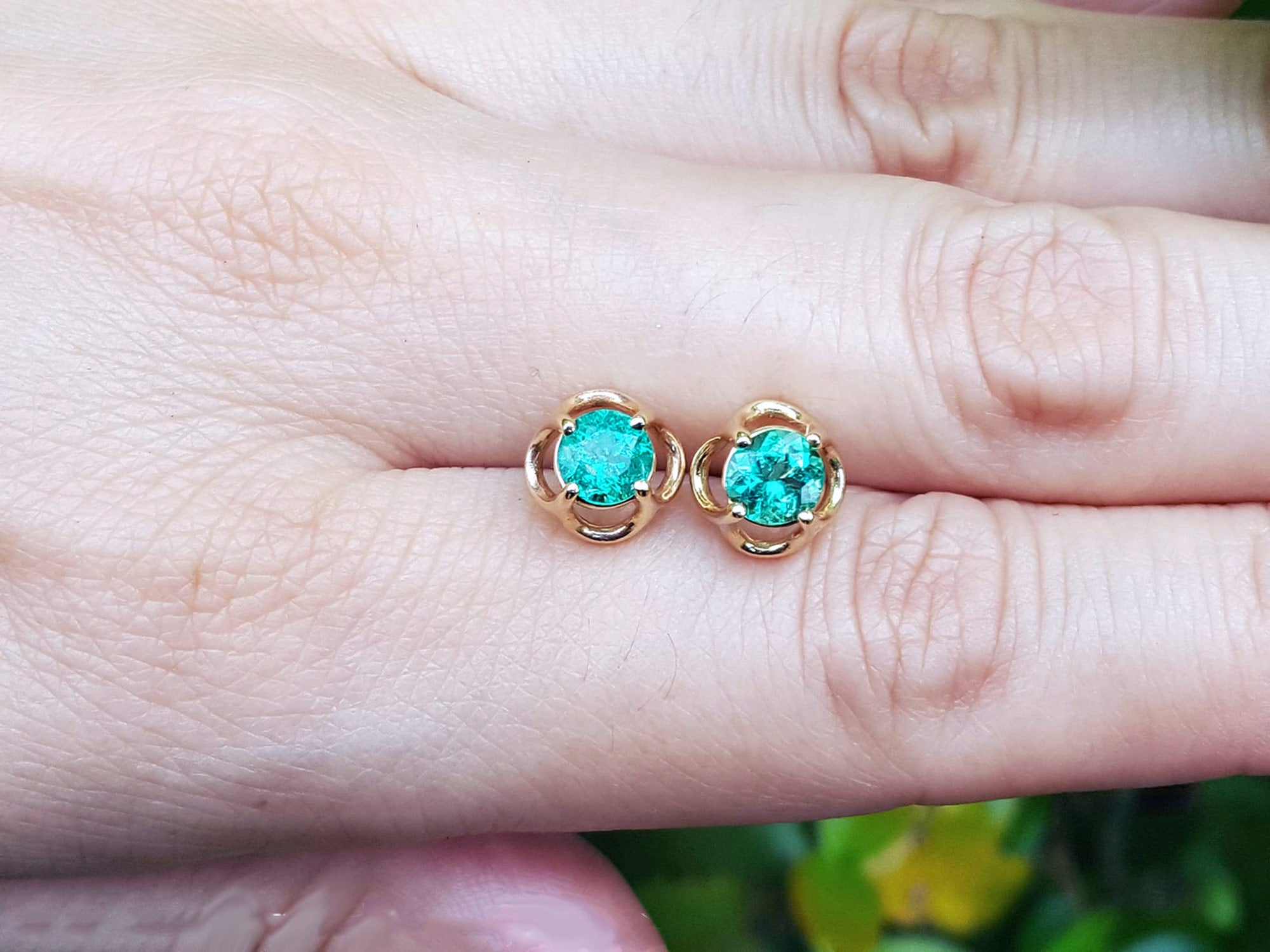 Yellow gold tulip emerald stud earrings held between fingers on a light skin background.