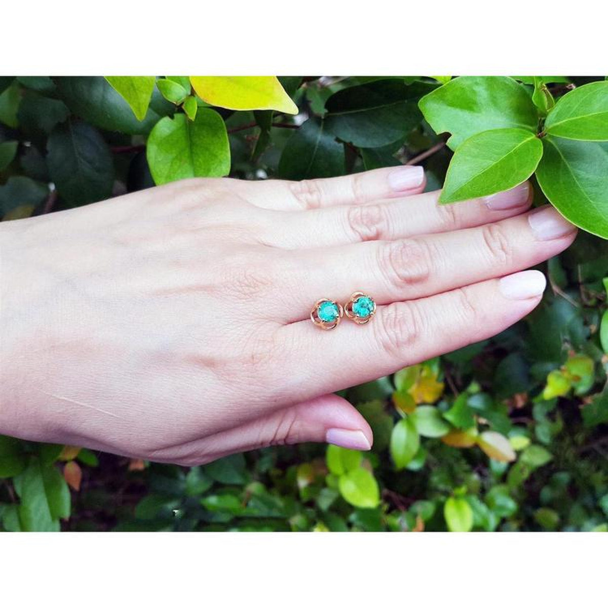 Colombian emerald tulip stud earrings in 14K yellow gold, worn on a hand against green foliage.