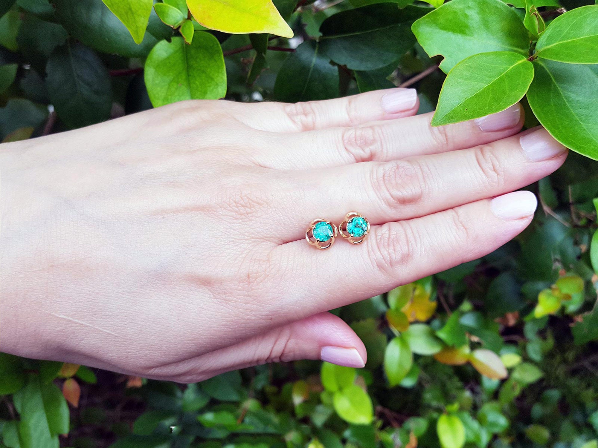 Hand displaying a pair of natural tulip emerald stud earrings against a green leafy background.