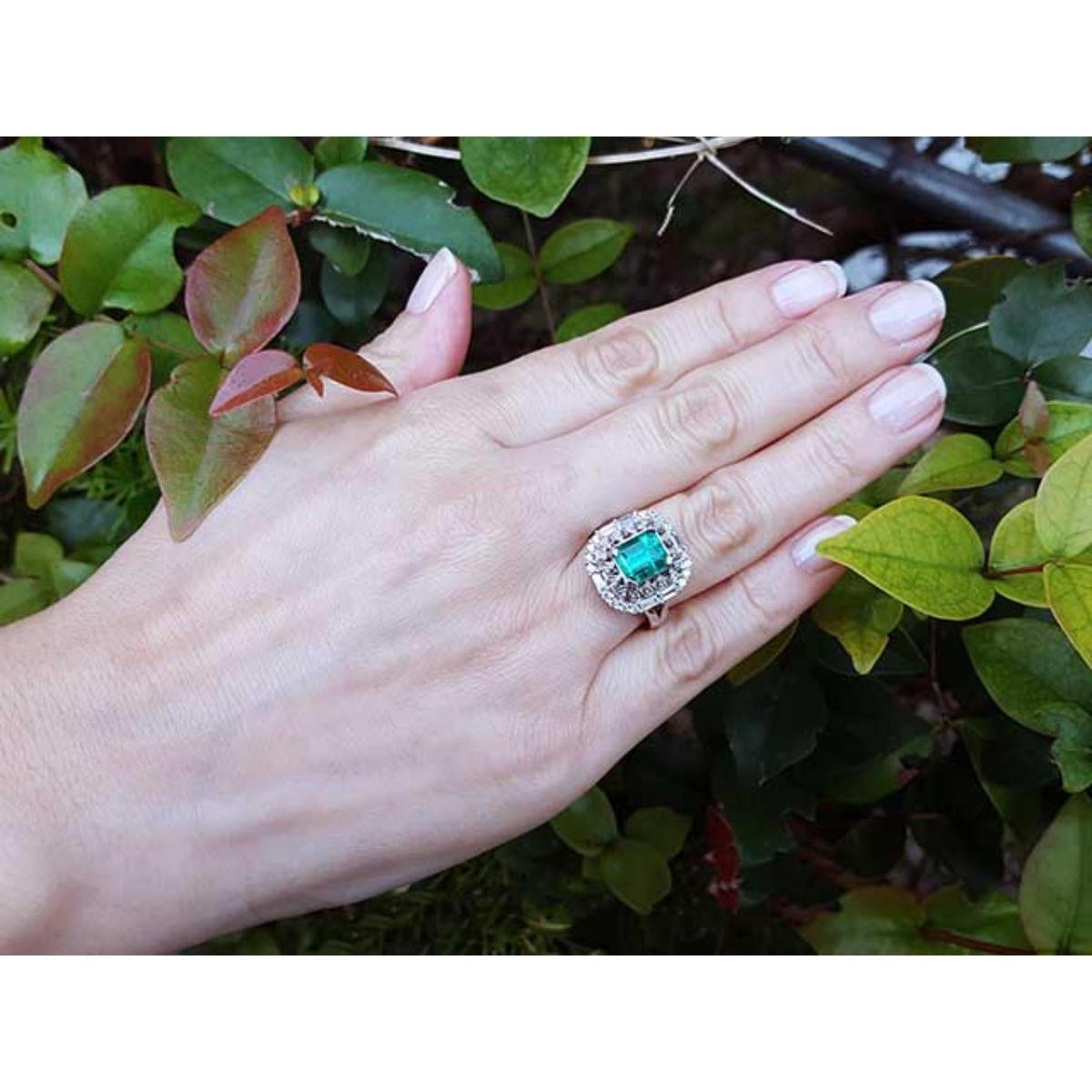 Hand wearing a vintage emerald and diamond ring against a green leafy background.