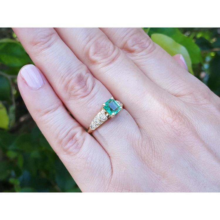 A square natural colombian emerald ring on a hand against a blurred green background.