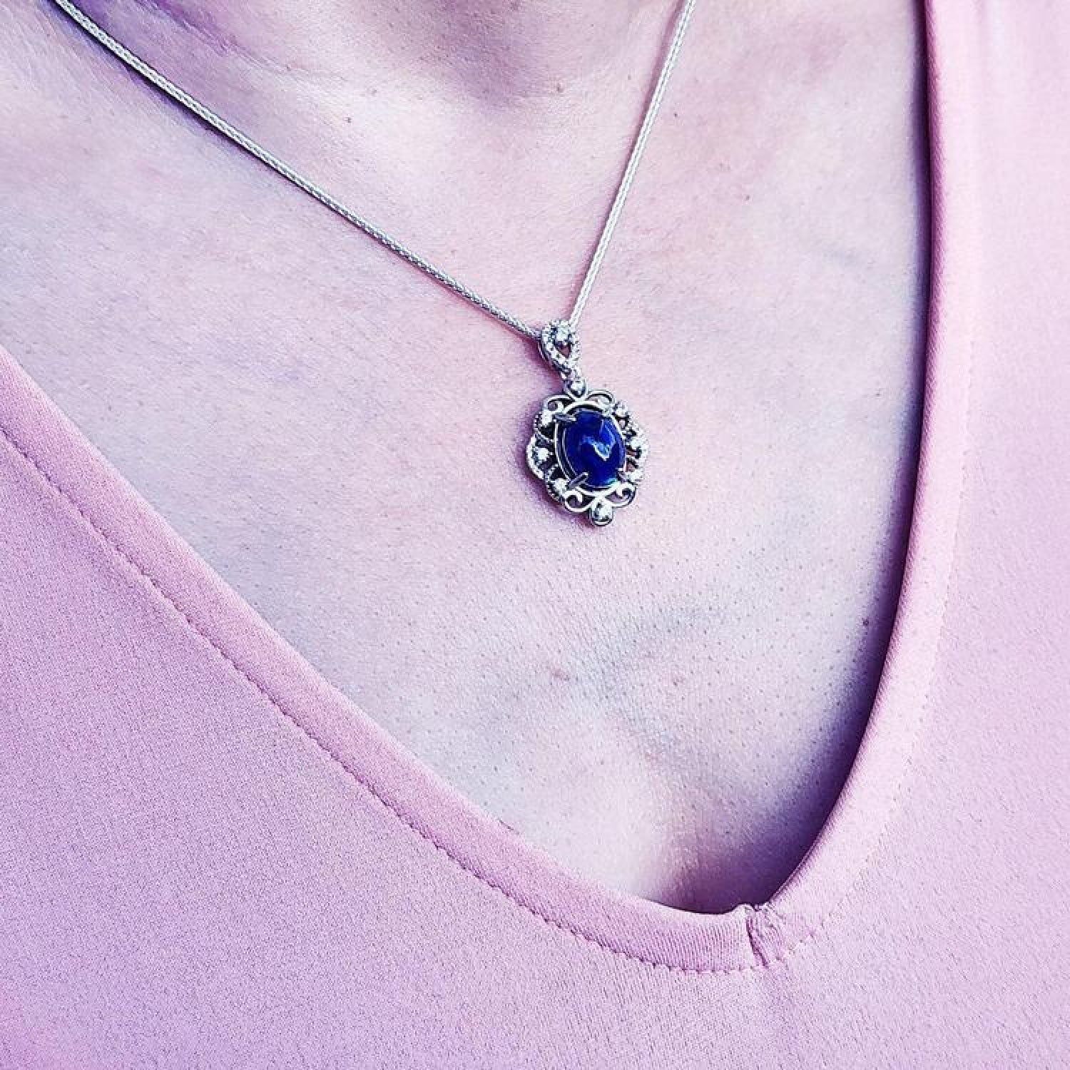Silver pendant with an oval, deep blue gemstone, surrounded by intricate silver scrollwork, worn on a delicate chain.