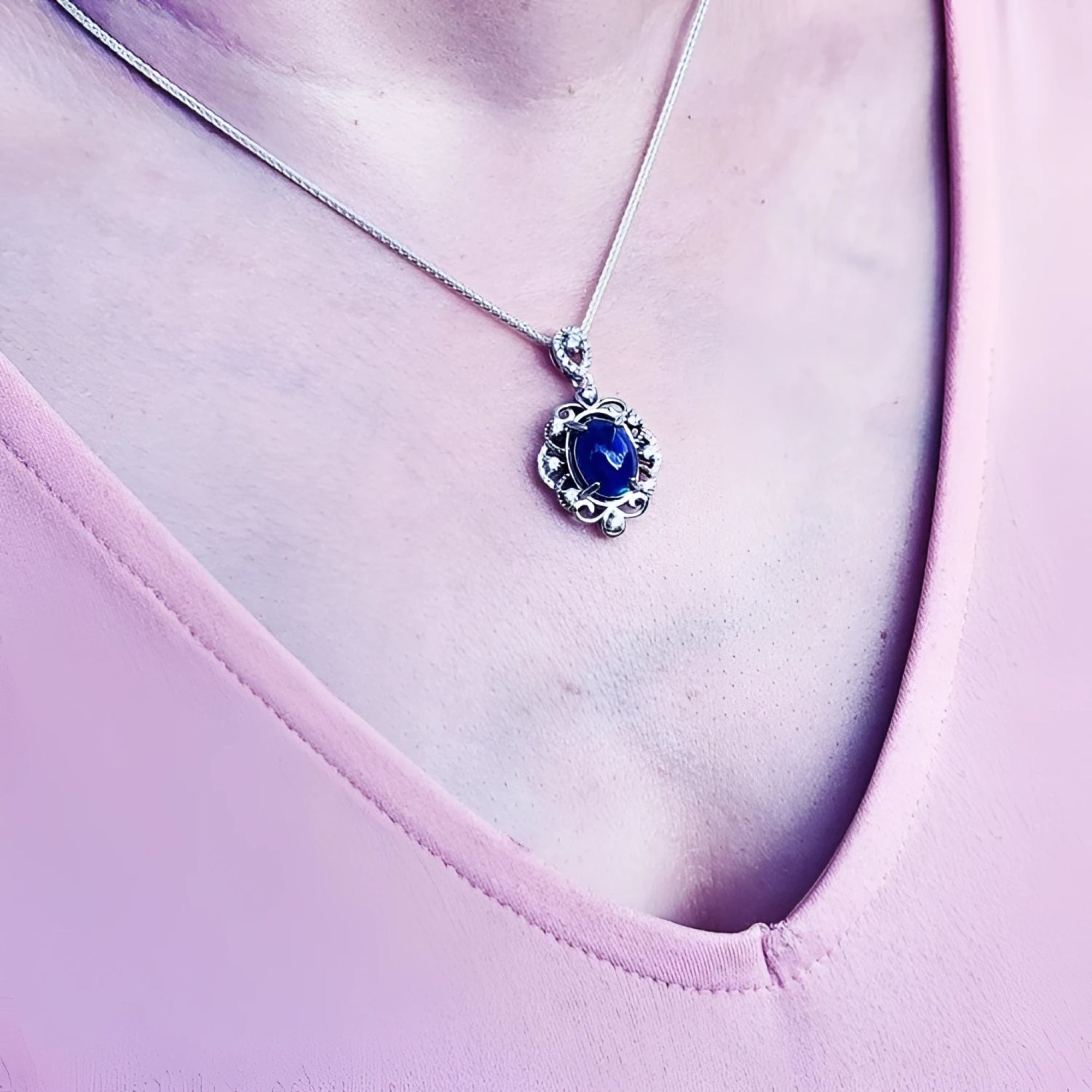Silver pendant with an oval, deep blue gemstone, surrounded by intricate silver scrollwork, worn on a delicate chain.