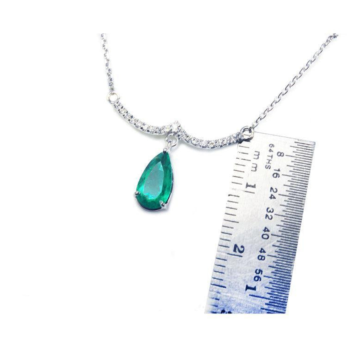 White gold necklace with a pear-shaped emerald and diamond-studded wavy bar, next to a ruler.
