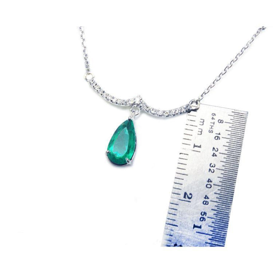 White gold necklace with a pear-shaped emerald and diamond-studded wavy bar, next to a ruler.