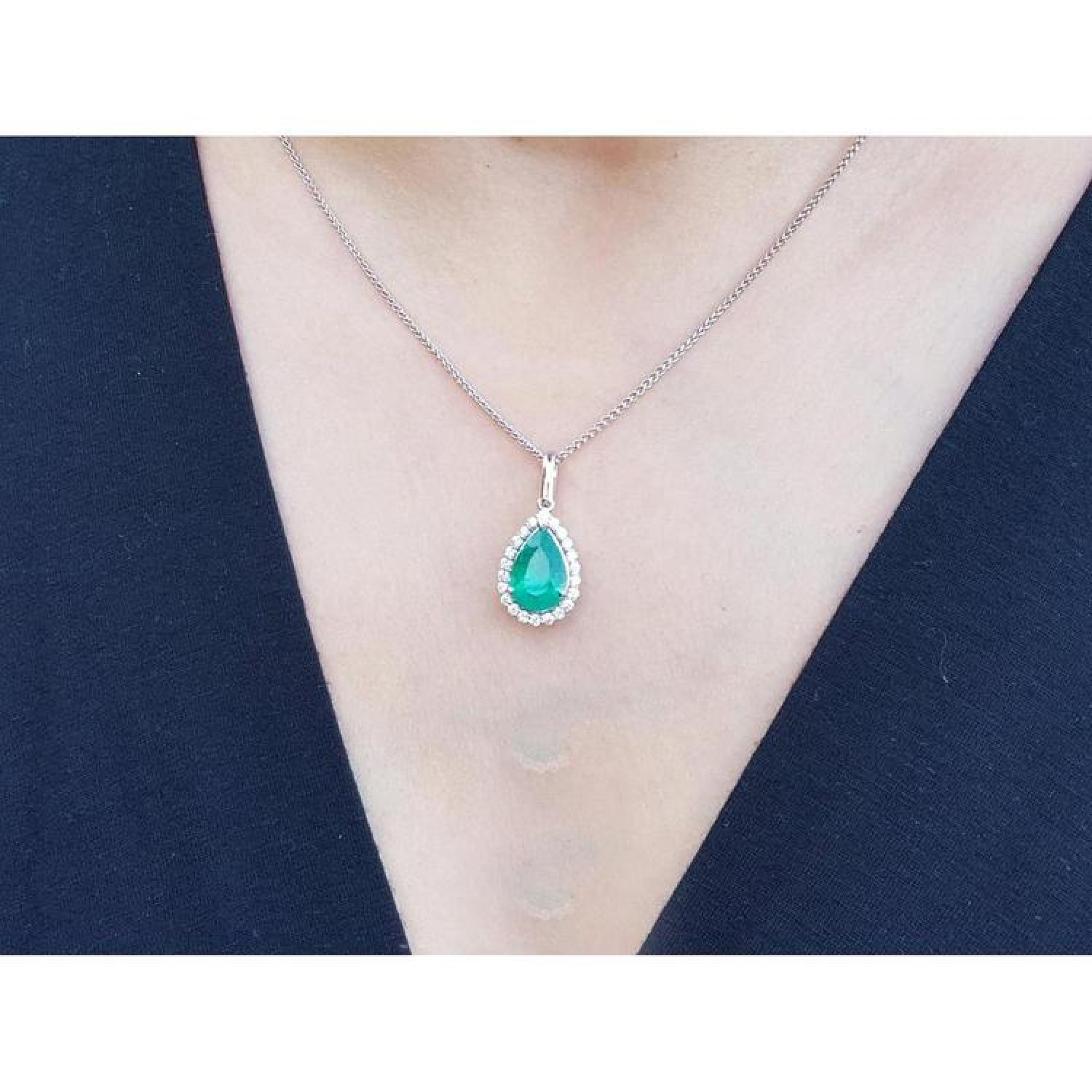 Pear-shaped deep green emerald pendant with a diamond halo on an 18K white gold chain, worn on a person.