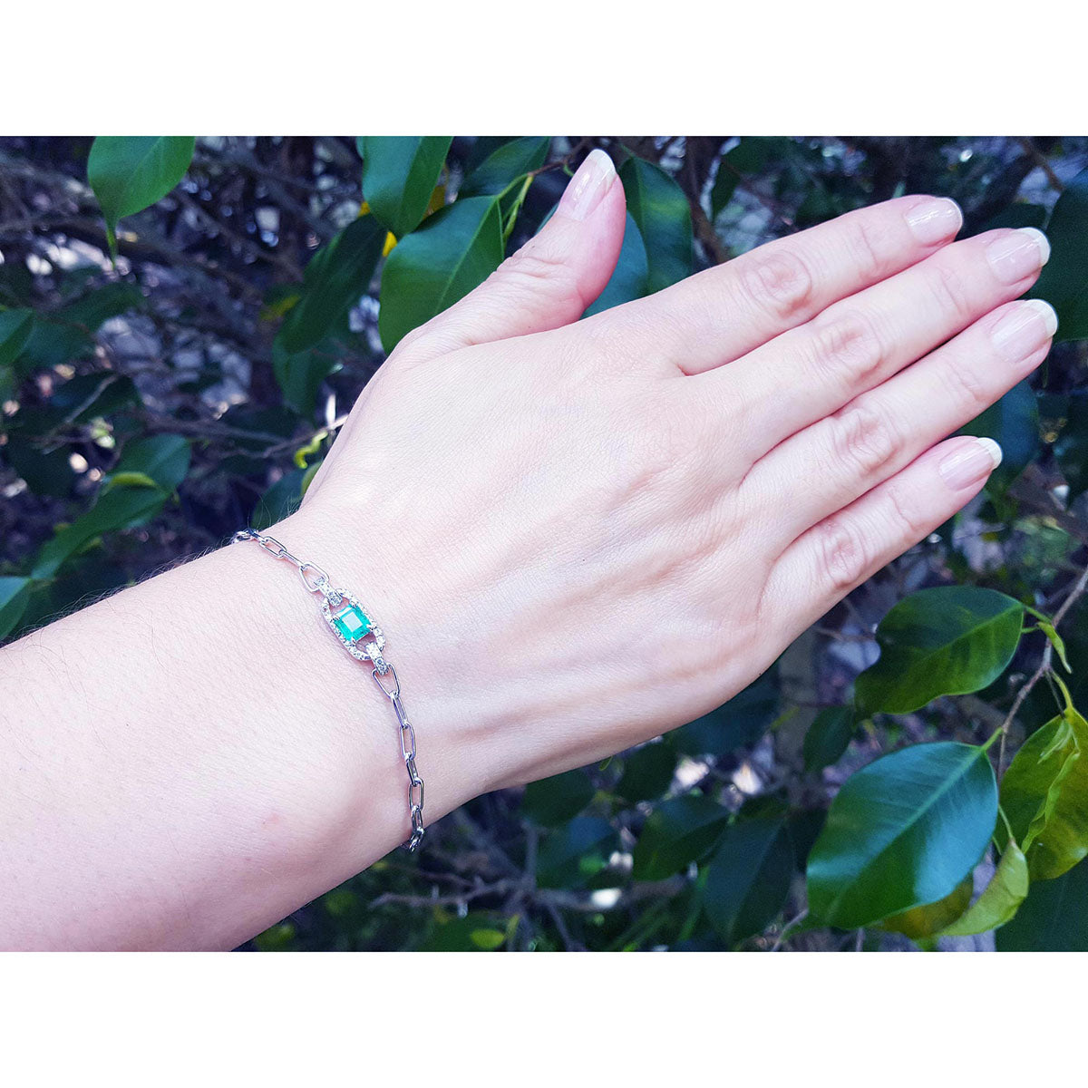 Hand wearing a white gold bracelet with a genuine emerald against a leafy background.