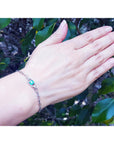 Hand wearing a white gold bracelet with a genuine emerald against a leafy background.