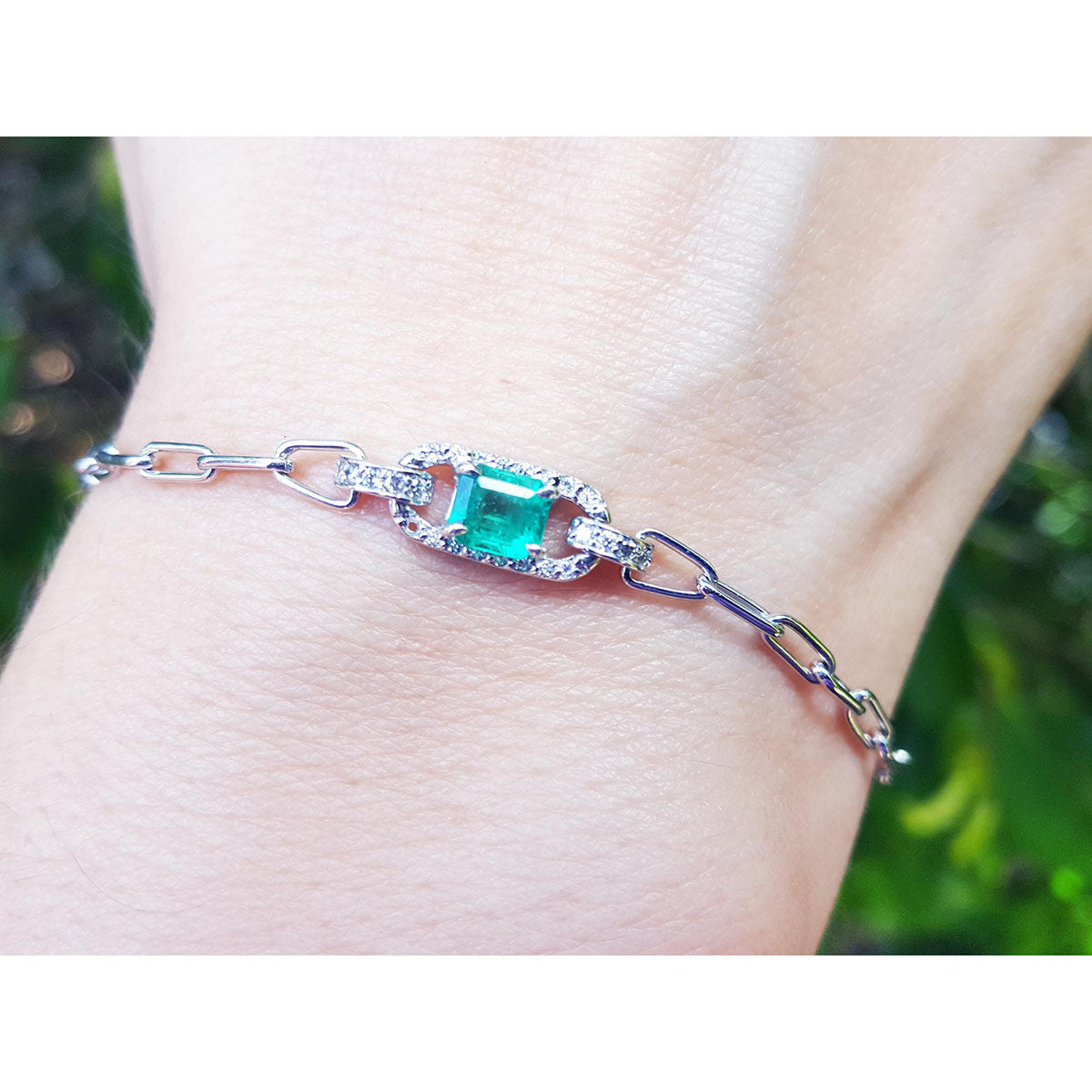 A white gold paperclip bracelet with a square emerald on a wrist against a blurred natural background.