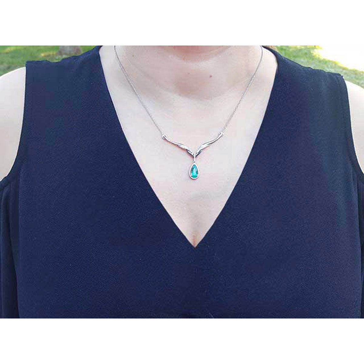 A lady wearing a blue dress with a white gold pear cut emerald necklace featuring a halo diamond pendant.