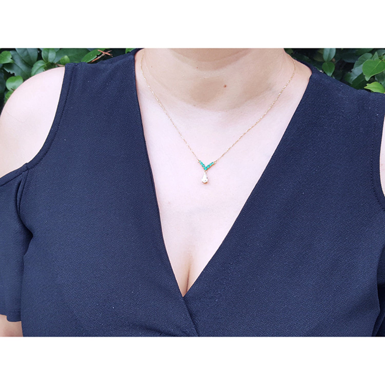 A close-up of a person wearing a 14K gold necklace with a 0.69 ct pear diamond and 0.35 ct round emeralds on a woman's neck., styled with a dark navy cold-shoulder top.