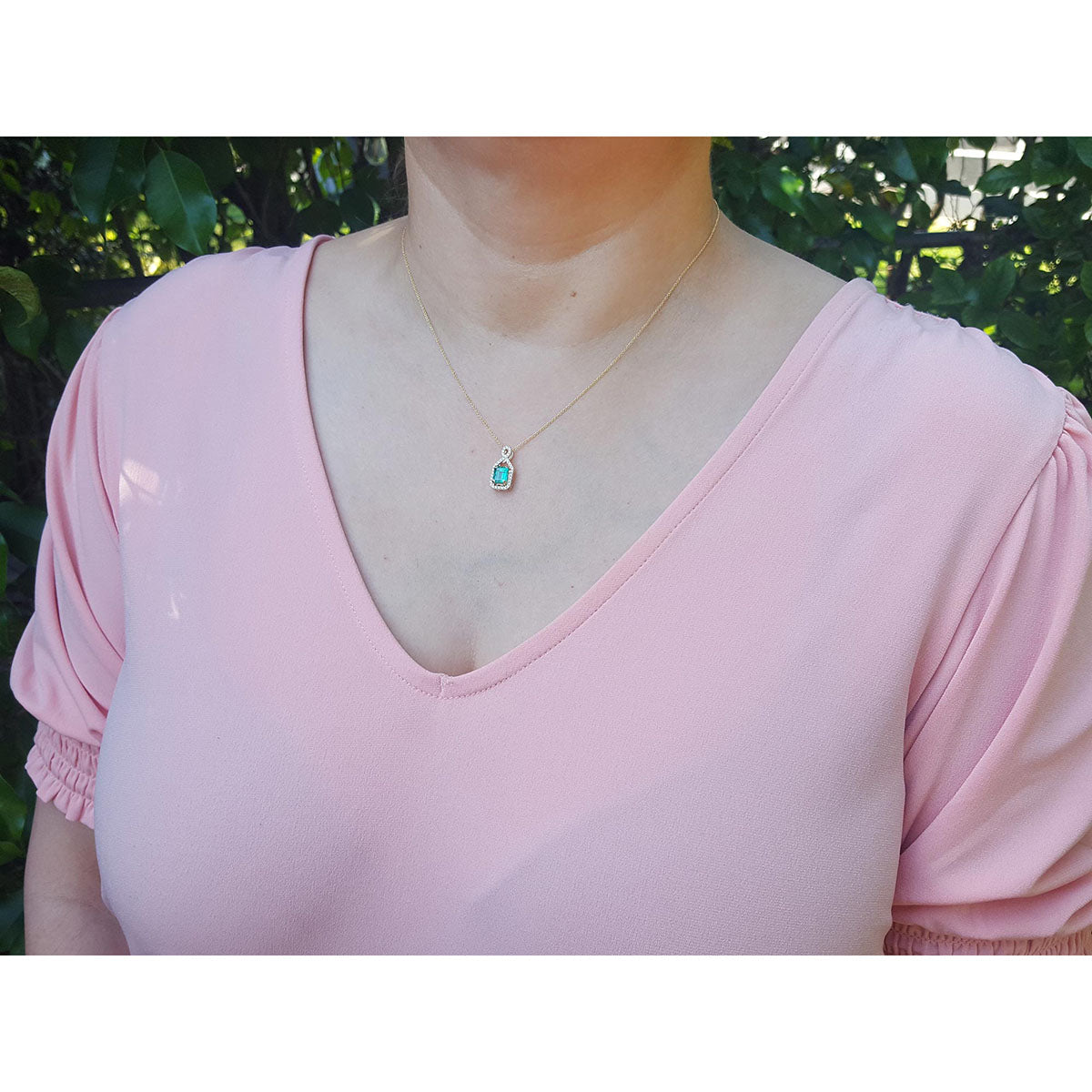 Woman wearing a pink top with a necklace featuring an emerald pendant against a blurred natural background