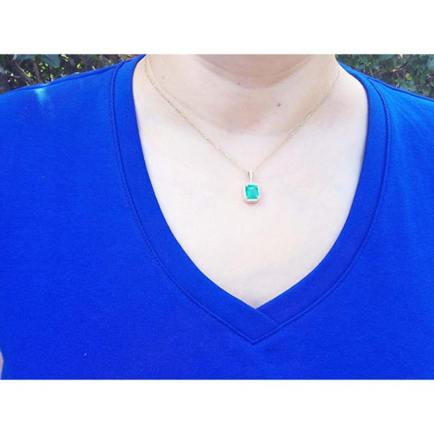 Emerald-cut Colombian emerald pendant with diamond halo on 14k yellow gold chain, worn on a blue shirt.