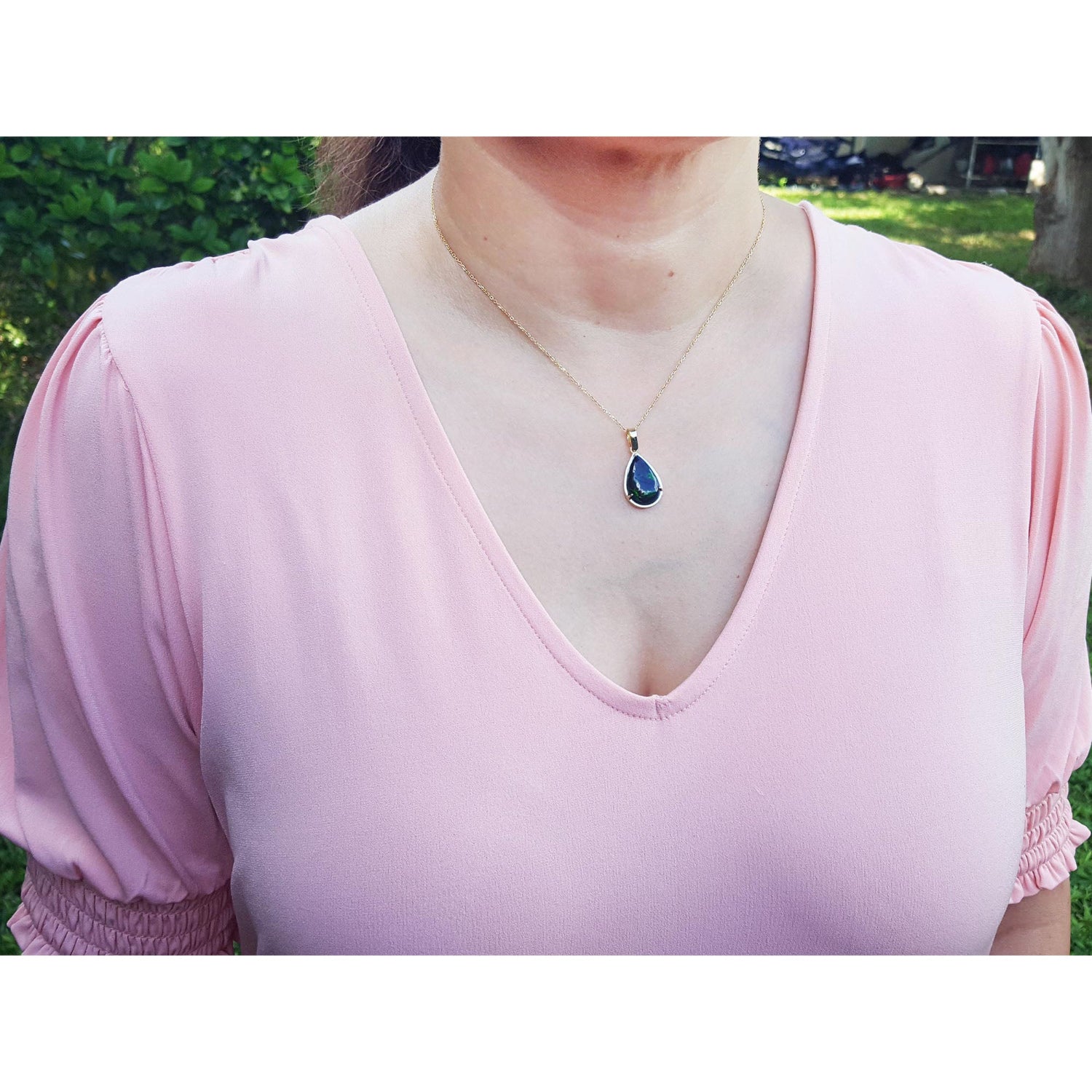 Person wearing a pink blouse with a necklace featuring an Ethiopian black opal gold pendant, outdoors.