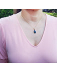 Person wearing a pink blouse with a necklace featuring an Ethiopian black opal gold pendant, outdoors.