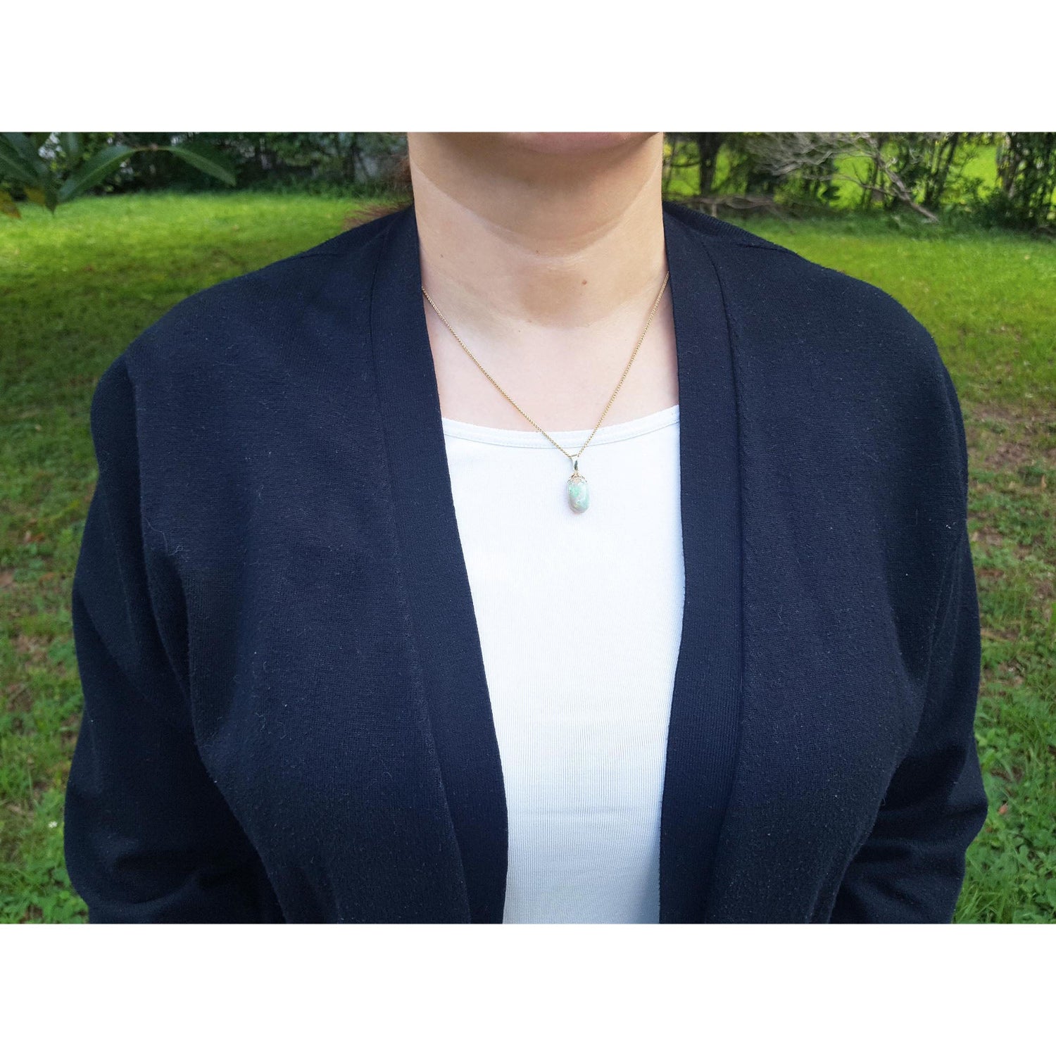 Person wearing a navy cardigan over a white shirt with an Ethiopian opal pendant on a blurred green outdoor background.