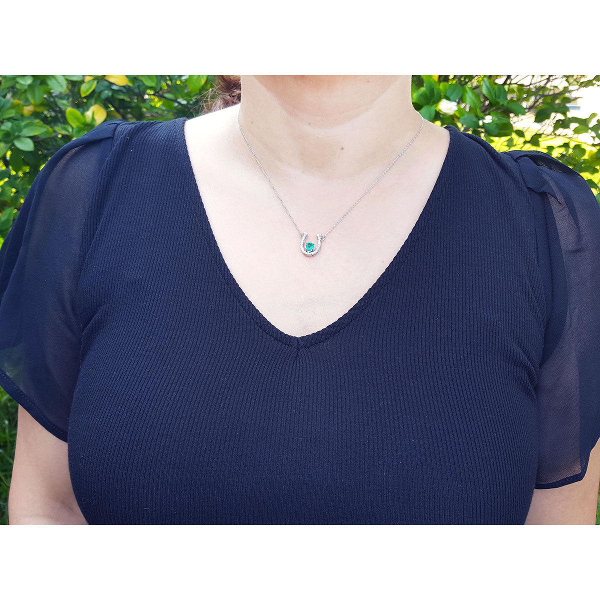 Woman wearing a navy blue top with a horseshoe emerald necklace against a green outdoor background