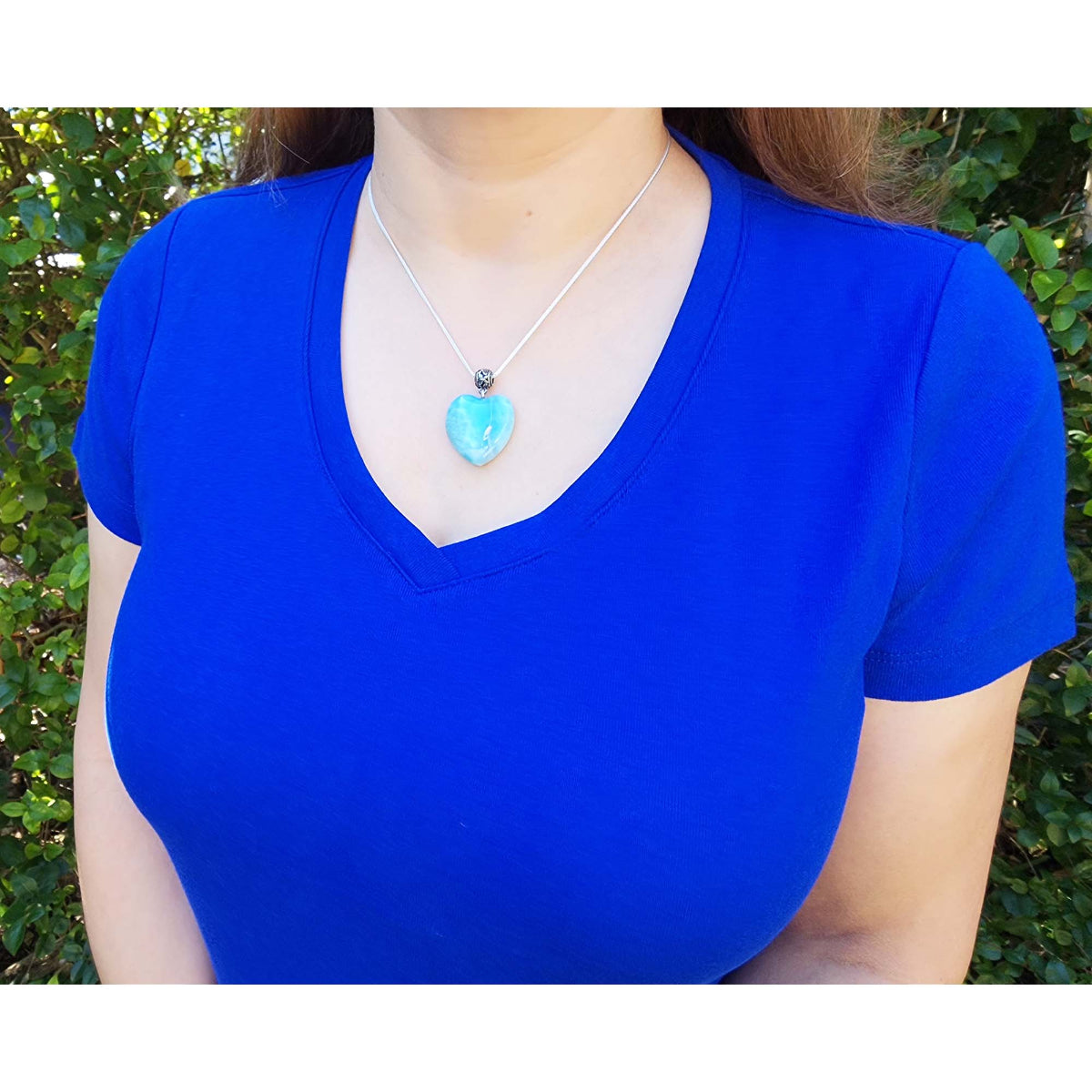 Person wearing a blue shirt with a necklace featuring a heart-shaped larimar pendant outdoors.