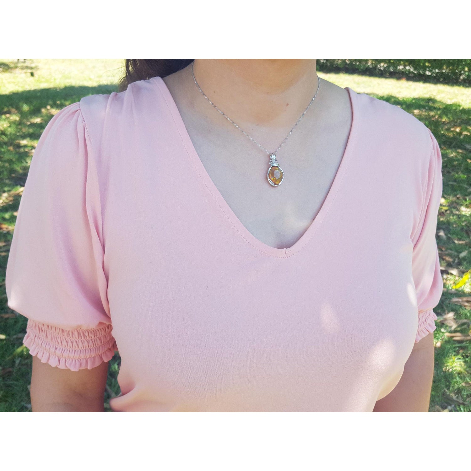 A woman wearing a pink blouse with ruffled sleeves and an opal silver pendant necklace outdoors.