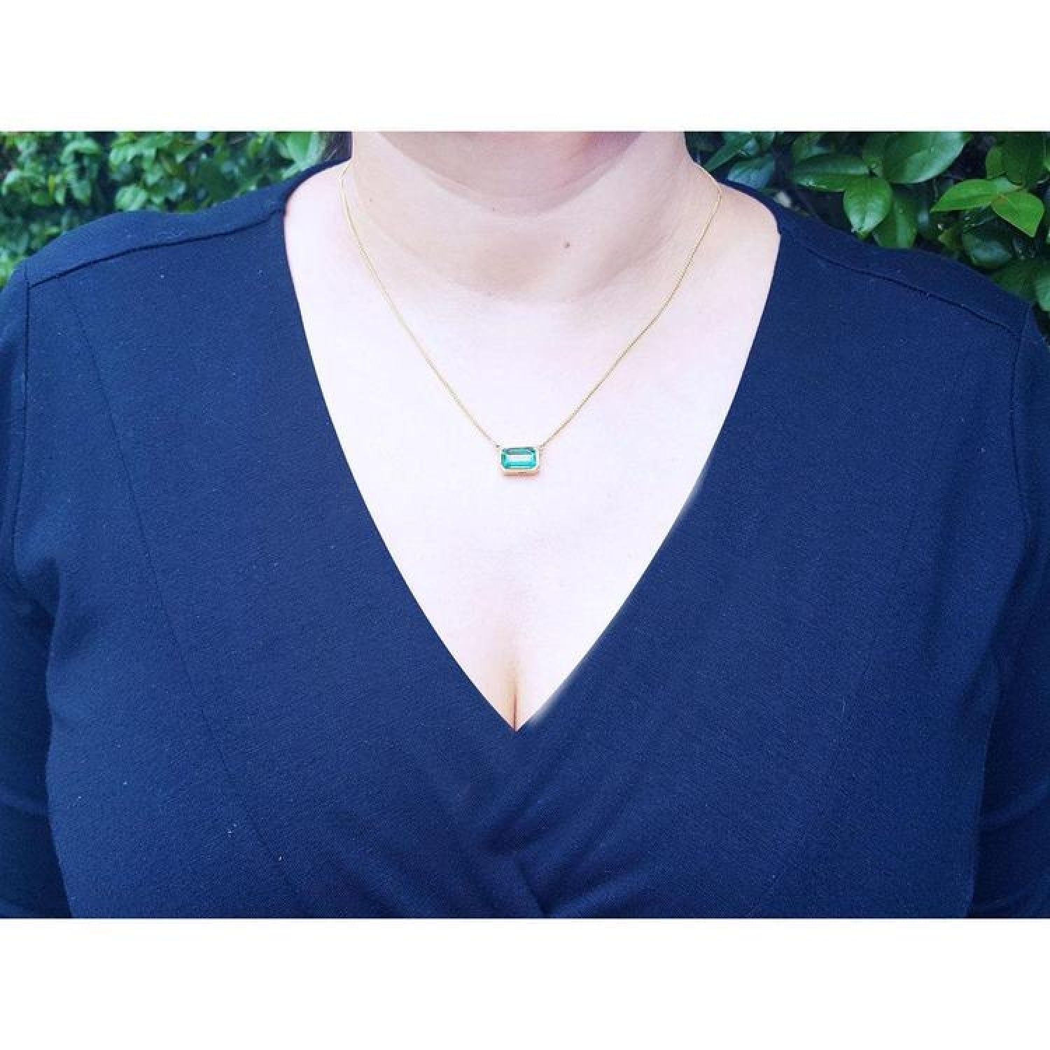 Woman wearing an 18K yellow gold necklace with a bezel-set, east-west emerald-cut bluish-green emerald.
