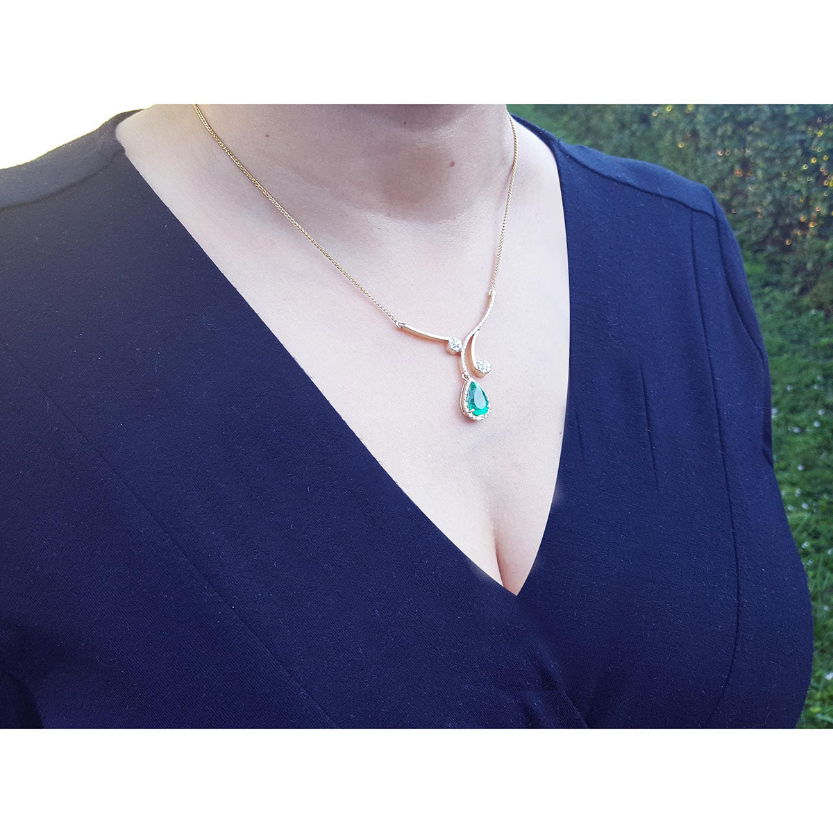 woman wearing a blue dress with a gold necklace featuring a teardrop emerald halo diamond pendant.