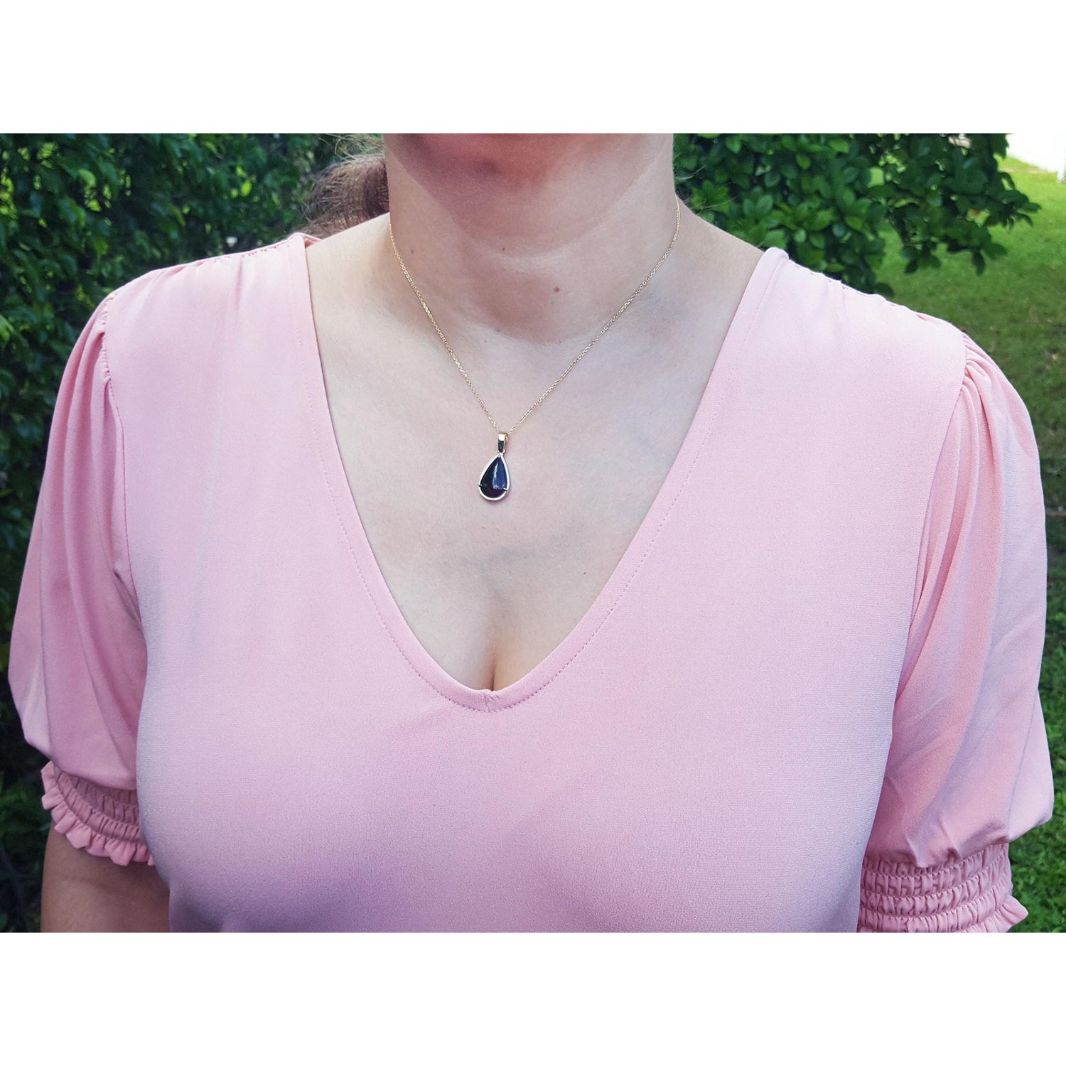 Person wearing a pink blouse with an opal necklace against a green outdoor background.