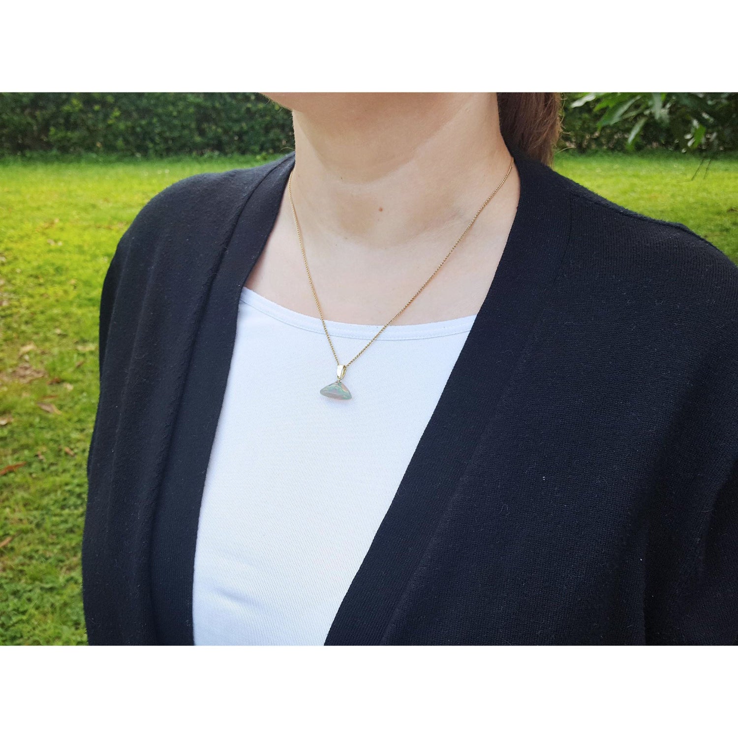 Person wearing a black cardigan over a white shirt with a necklace featuring an opal pendant outdoors.