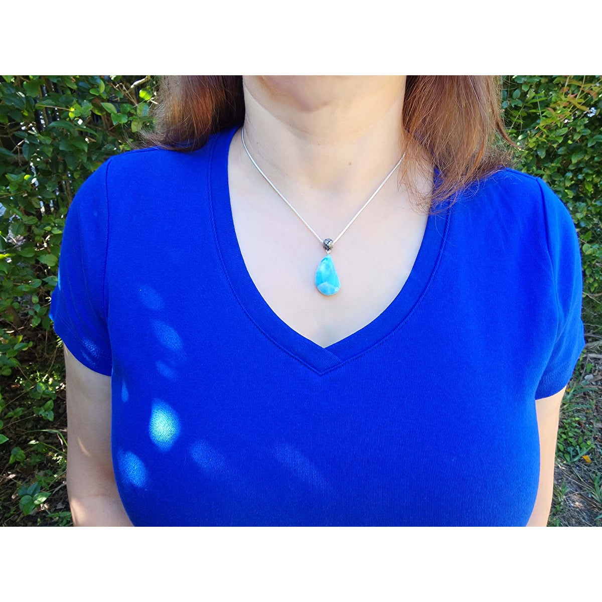 Person wearing a blue shirt with a necklace featuring a larimar pendant against a green outdoor background.