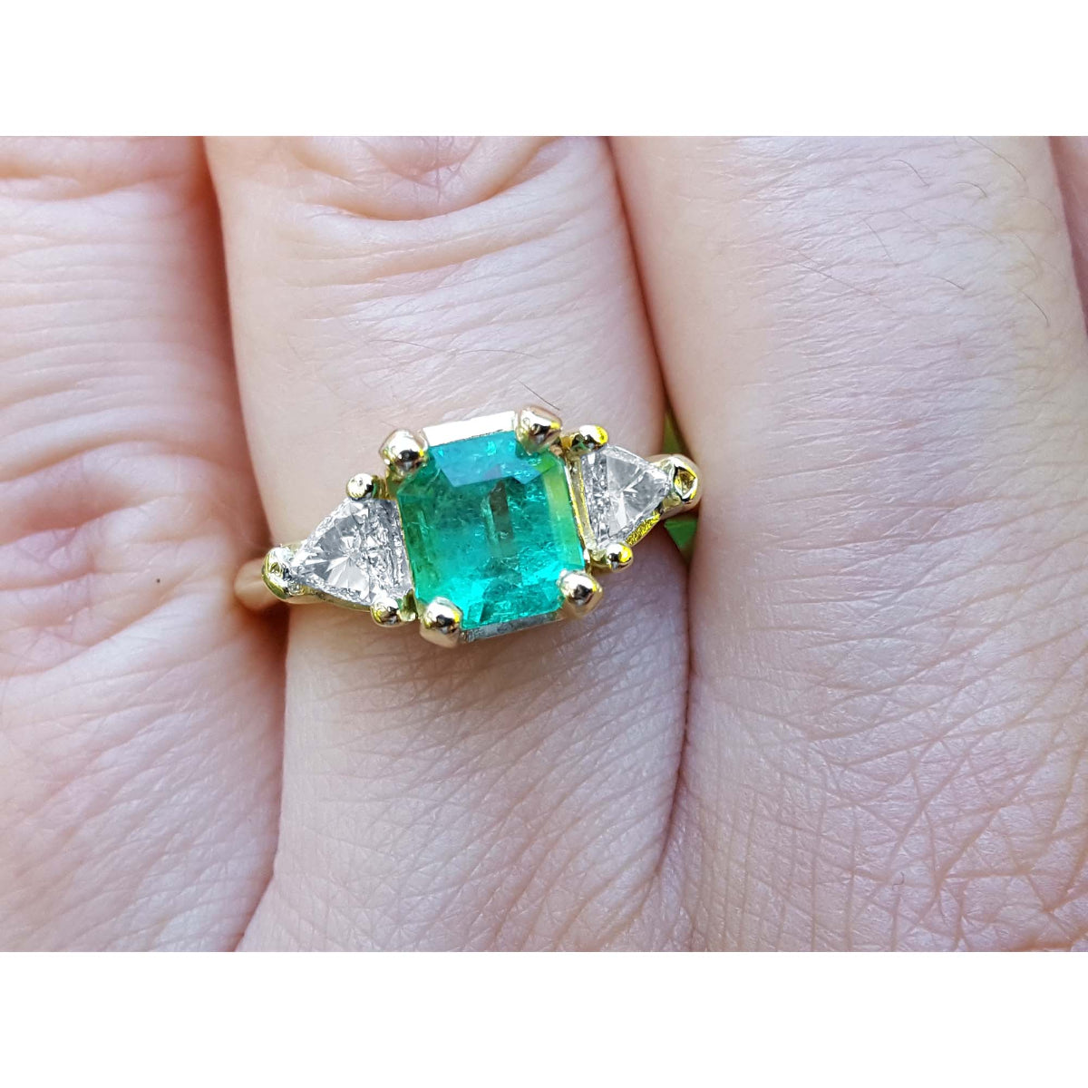 Square emerald and trillion side diamonds trillion cut ring made of 18k yellow gold on a finger.
