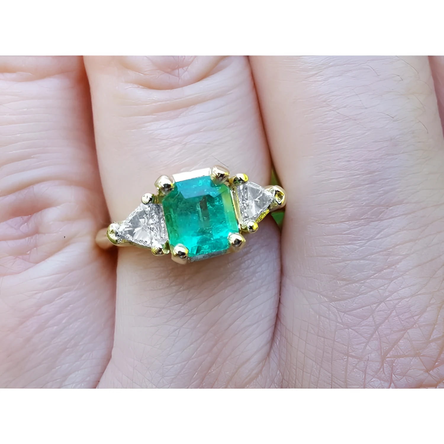 Square emerald and trillion side diamonds trillion cut ring made of 18k yellow gold on a finger.