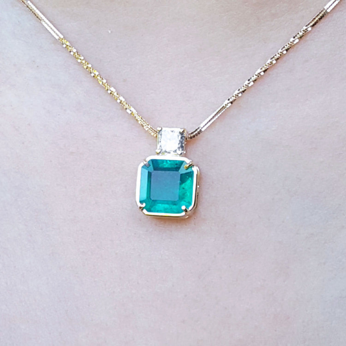 Front side of a yellow gold necklace with an Asscher cut emerald gemstone pendant on a light background