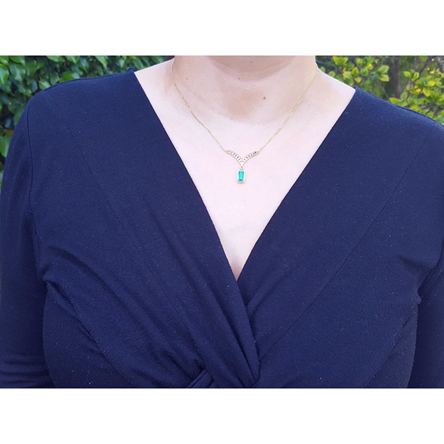 A close-up of a person wearing a 14K yellow gold necklace with a baguette-cut Colombian emerald pendant dangling from a diamond-studded V, paired with a navy-blue wrap top.