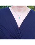 A close-up of a person wearing a 14K yellow gold necklace with a baguette-cut Colombian emerald pendant dangling from a diamond-studded V, paired with a navy-blue wrap top.