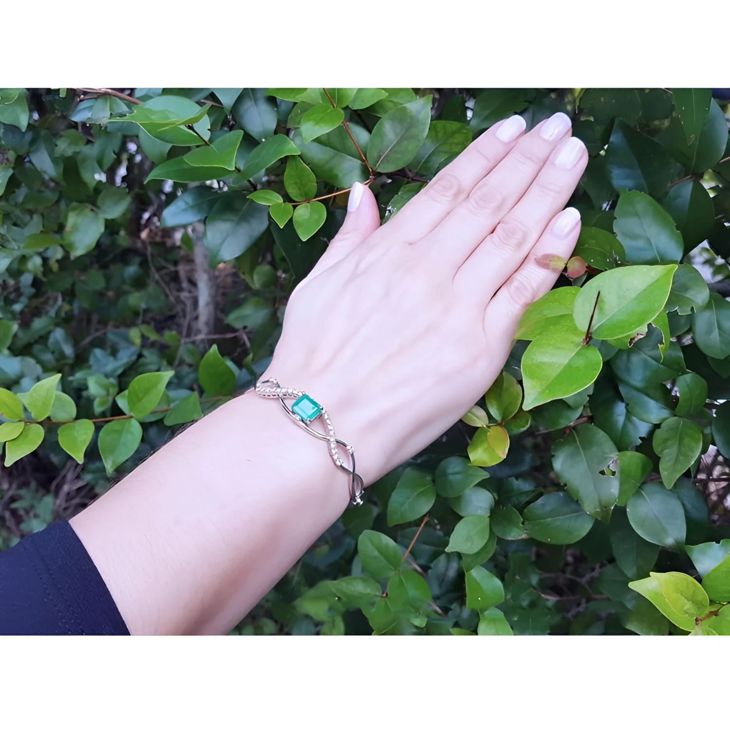 Emerald bangle bracelet with natural diamonds on 14K white gold, worn on a hand against green leaves.