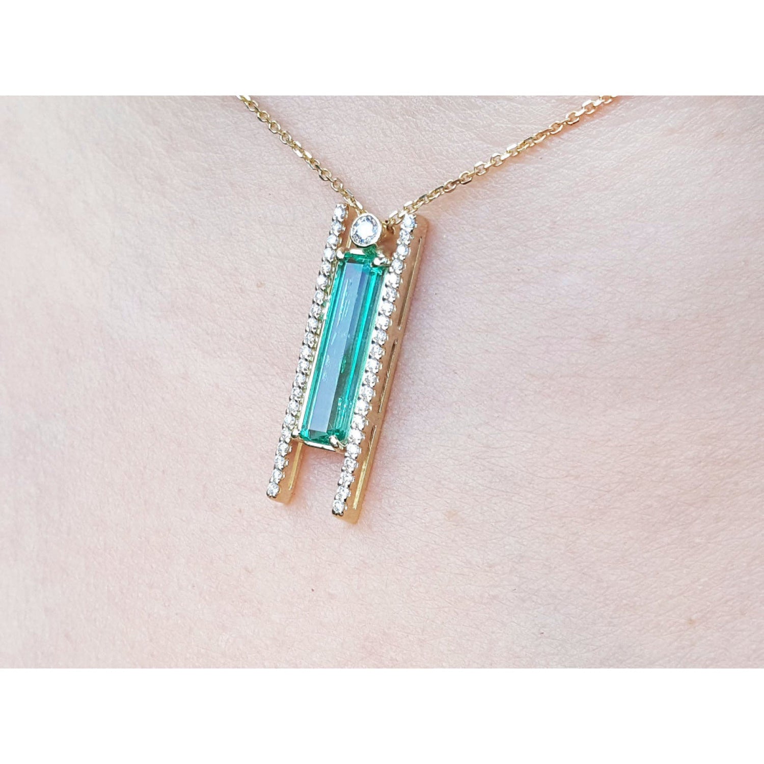 A close-up of a 14K yellow gold necklace with a baguette-cut emerald and natural diamond accents worn on a person's neck.