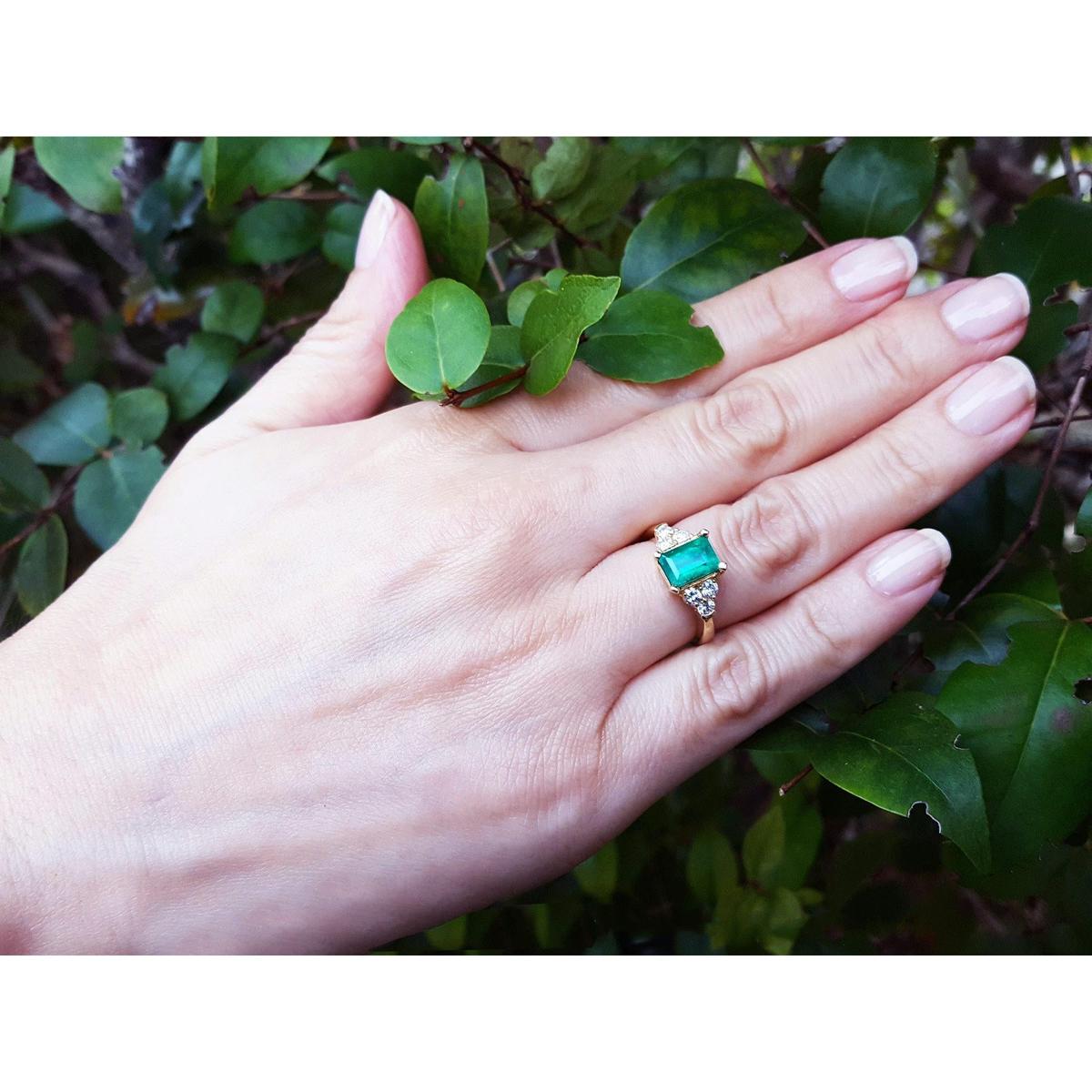 Hand wearing a yellow gold emerald ring with a leafy background.