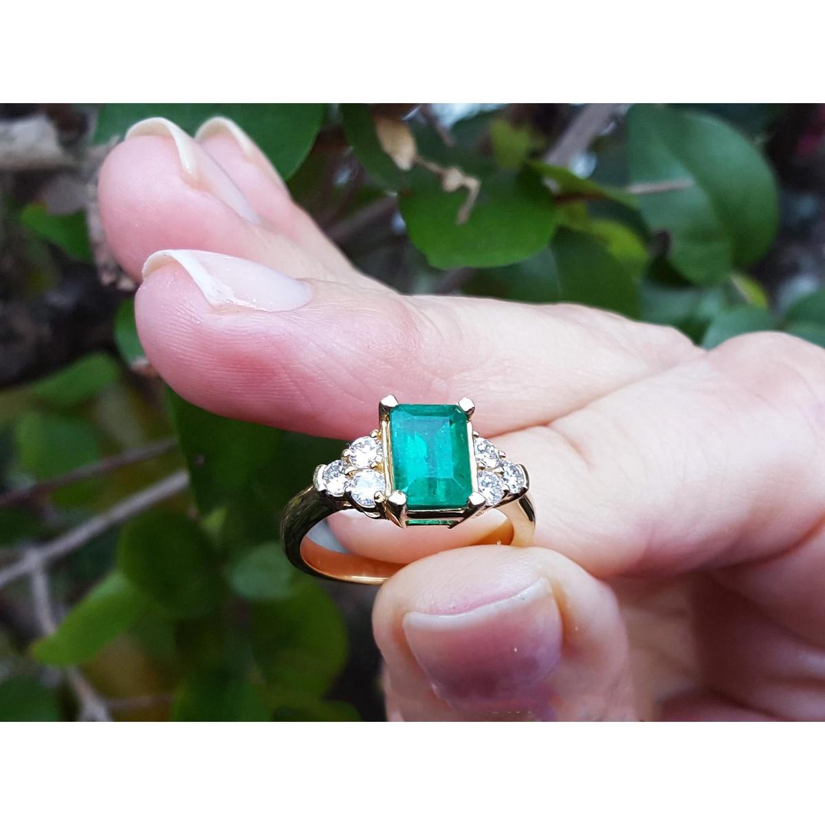 A yellow gold ring with emerald and side diamonds held between fingers.