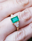 A yellow gold ring with a natural emerald and diamond accents on a finger.