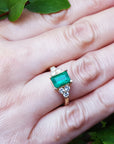 Gold ring with an emerald and diamond accents on a leafy background.