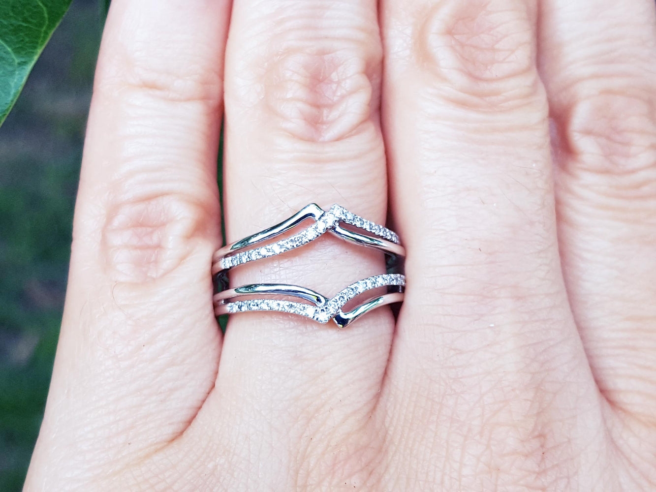 Close-up of a hand wearing a diamond ring guard enhancer wrapper 14k white gold with a blurred green background.